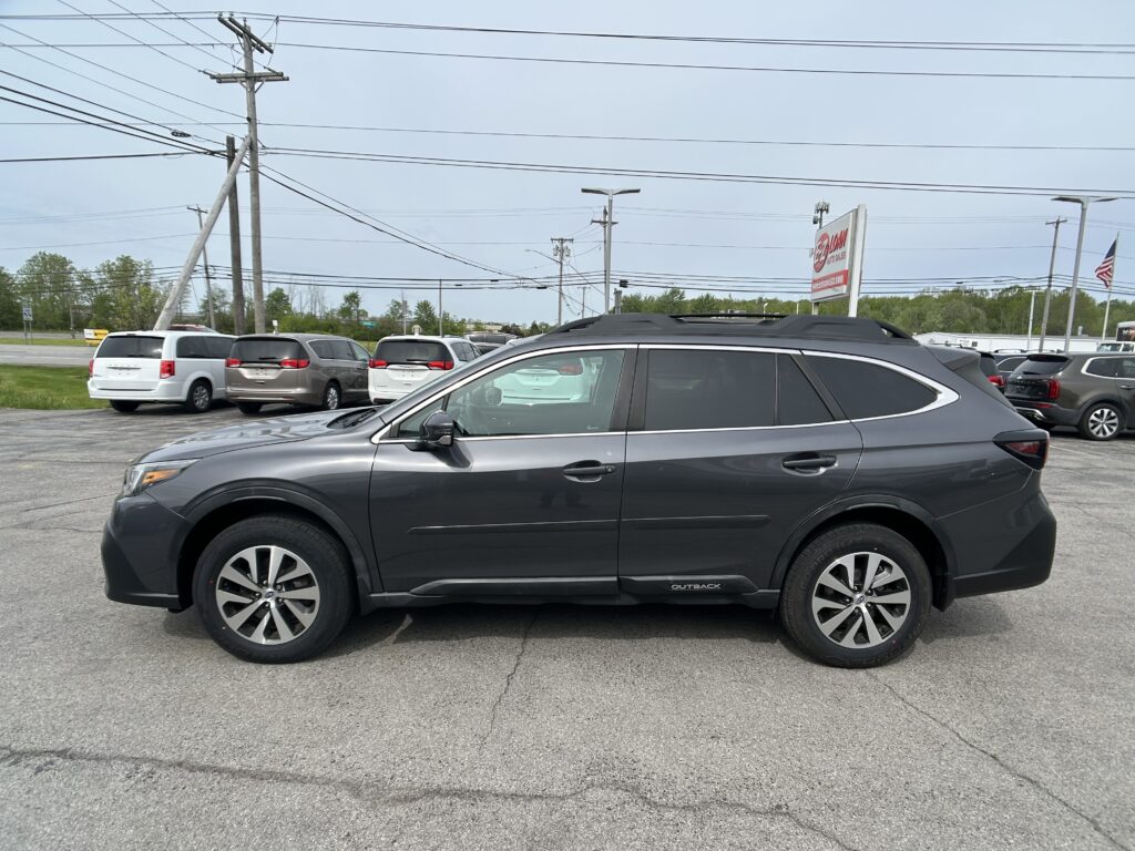 Side view of a gray Subaru Outback SUV parked in a busy car lot with other minivans and power lines overhead.