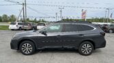 Side view of a gray Subaru Outback SUV parked in a busy car lot with other minivans and power lines overhead.