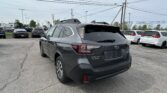 Rear view of a gray Subaru SUV in a crowded car dealership lot with power poles and overhead wires in the background.