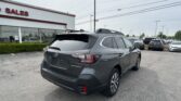Gray Subaru Outback SUV parked in a dealership lot with a white building labeled 'SALES' and numerous cars in the background.