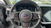 Close-up view of a car steering wheel inside a Subaru, with the instrument cluster and a right-side touchscreen visible, and a parking lot seen through the windshield.