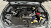 Open car hood revealing the engine bay with hoses, wires, and battery visible under the front of the vehicle.