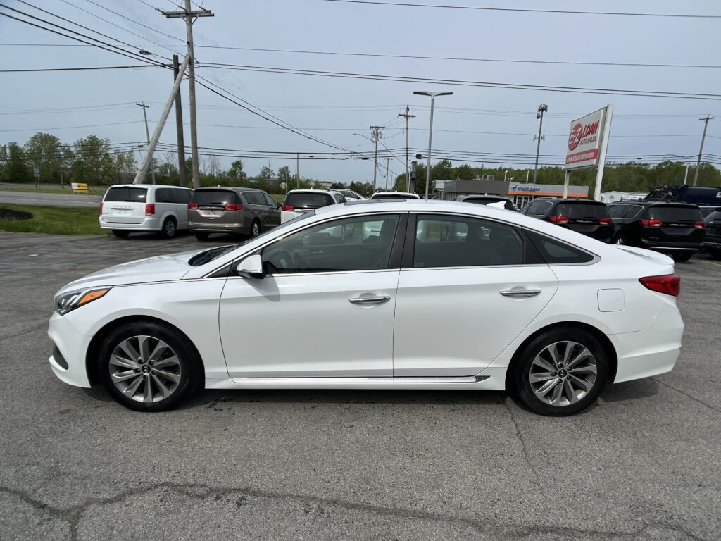 White sedan in foreground at a car dealership lot, with several minivans parked behind and power lines overhead.