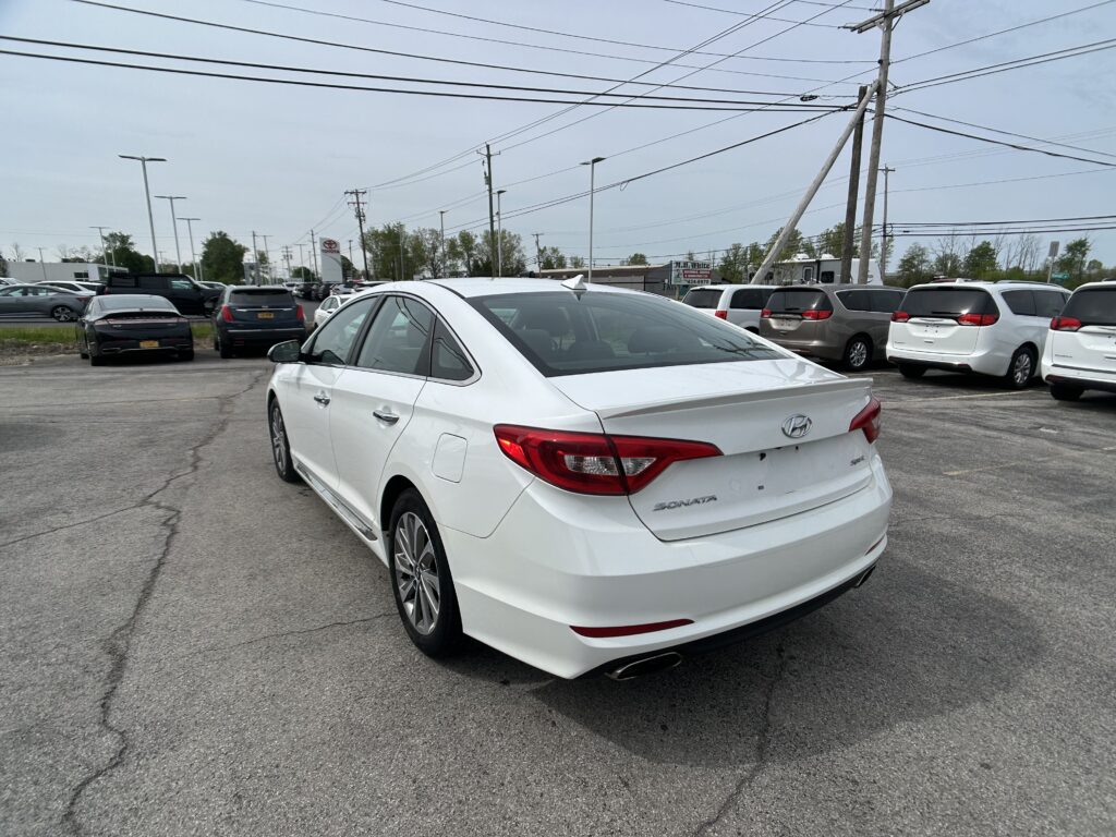 White Hyundai Sonata seen from the rear three-quarter view in a busy car dealership lot with many cars and overhead power lines in the background.