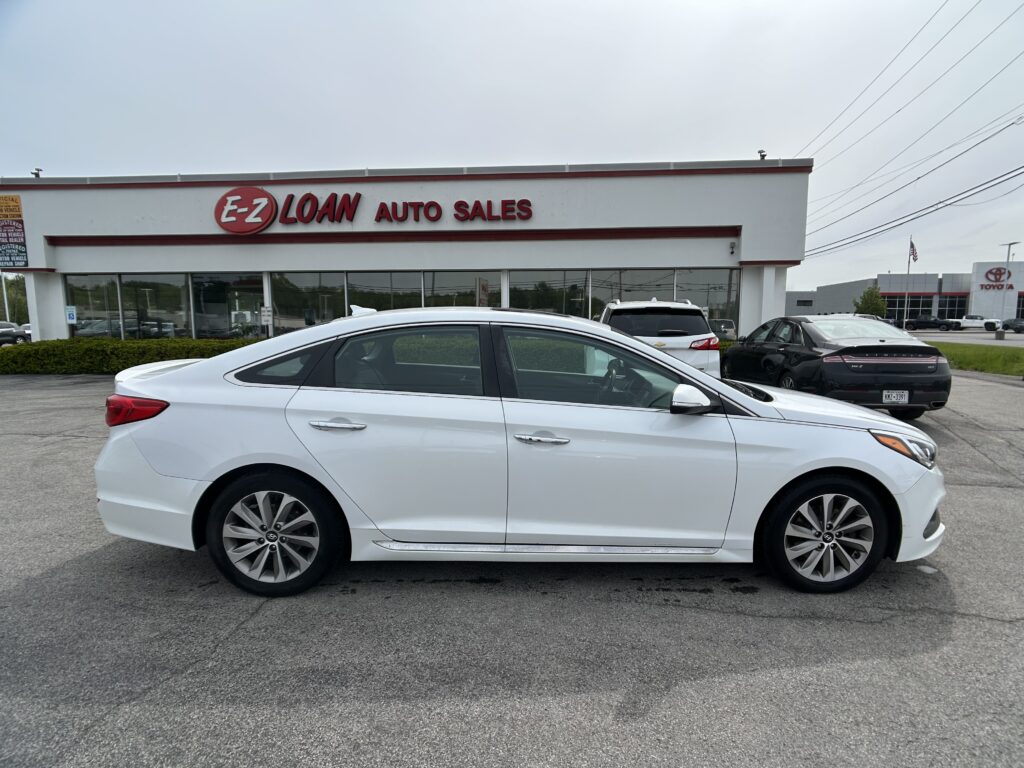 White sedan parked in the dealership lot in front of an E-Z Loan Auto Sales building, with other cars and power lines in the background.