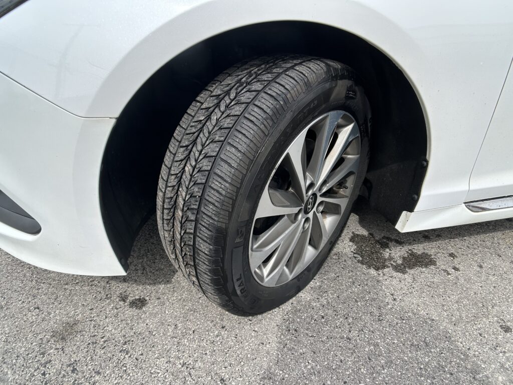 Close-up of a silver alloy wheel and treaded tire mounted on a white car, with the wheel well and fender visible on asphalt.