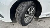 Close-up of a silver alloy wheel and treaded tire mounted on a white car, with the wheel well and fender visible on asphalt.