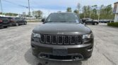 Front view of a gray Jeep SUV in a car dealership lot, with other vehicles and power lines in the background.