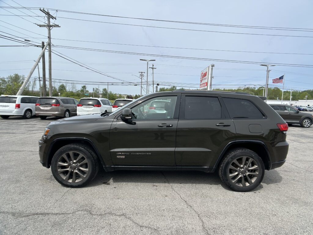 Close-up side view of a dark gray Jeep Grand Cherokee SUV parked in a dealership lot, with power lines and a clear blue sky behind.