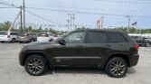 Close-up side view of a dark gray Jeep Grand Cherokee SUV parked in a dealership lot, with power lines and a clear blue sky behind.