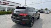 Back of a dark gray Jeep SUV in a car dealership lot with a white building that reads 'AUTO SALES' and rows of cars in the background under power lines.