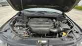 Open car hood view of a V6 engine bay with the battery on the left and various hoses and components visible beneath a black hood liner, in a parking lot.