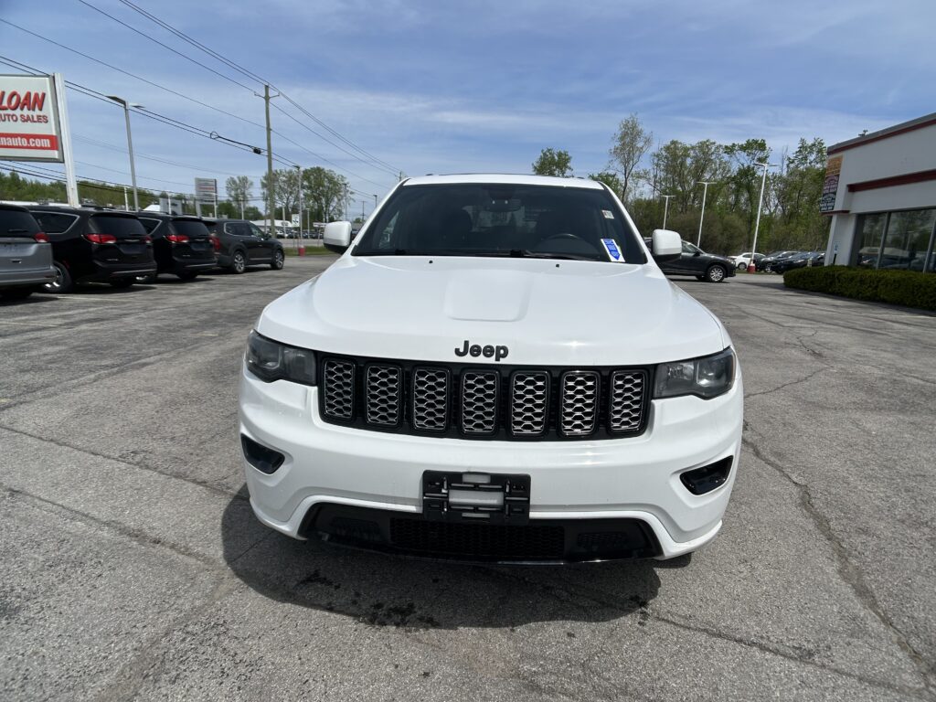 Front view of a white Jeep parked in a car dealership lot, with other vehicles and a dealership sign in the background on a clear day.