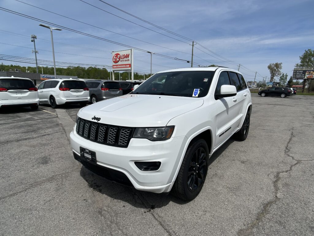 White Jeep SUV in a car dealership lot with other vehicles and a roadside EZ Loan sign in the background under a blue sky.