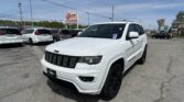 White Jeep SUV in a car dealership lot with other vehicles and a roadside EZ Loan sign in the background under a blue sky.