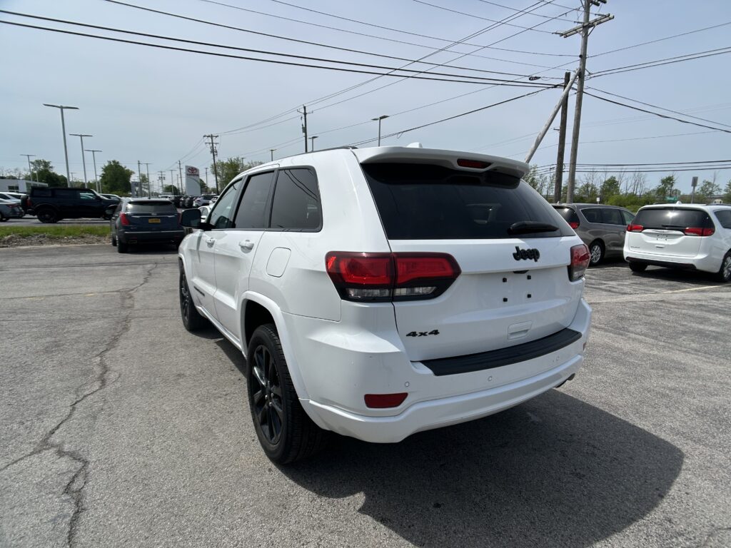 White Jeep SUV in the foreground at a car dealership lot, with other cars parked and tall power lines overhead against a clear sky.",