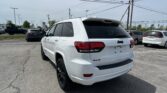 White Jeep SUV in the foreground at a car dealership lot, with other cars parked and tall power lines overhead against a clear sky.",