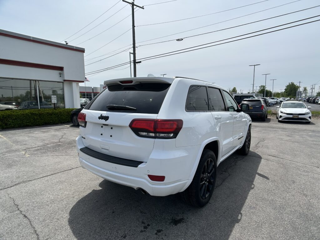 Rear three-quarter view of a white Jeep SUV parked in a car dealership lot with other vehicles and a building on the left.