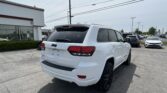 Rear three-quarter view of a white Jeep SUV parked in a car dealership lot with other vehicles and a building on the left.