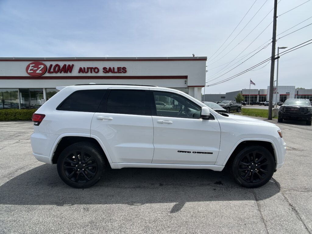 White Jeep Grand Cherokee SUV in the foreground of an auto dealership lot, with 'E-Z LOAN AUTO SALES' building in the background under a clear sky.