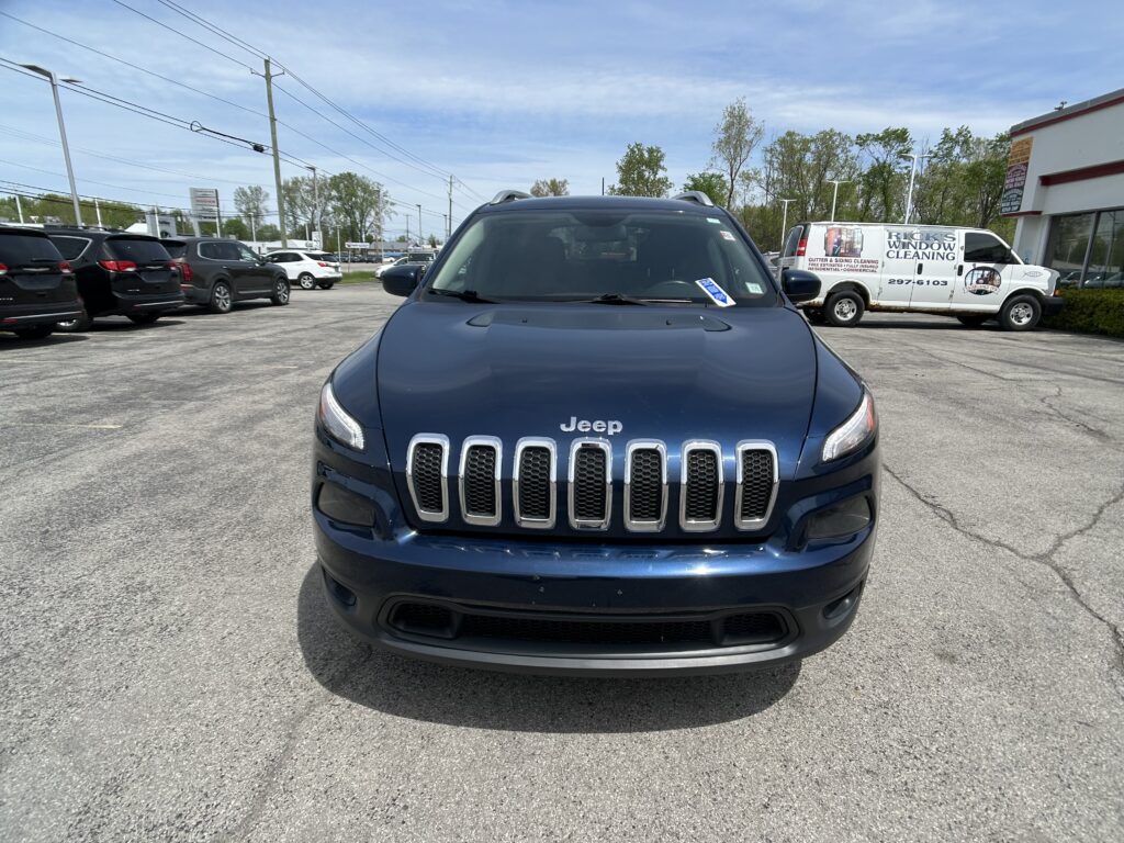 Front view of a blue Jeep SUV in a parking lot, with other cars and a white window-cleaning van in the background.