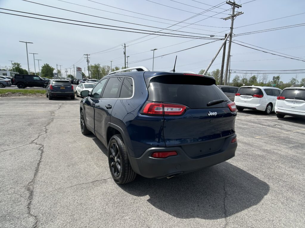 Blue Jeep SUV in a parking lot with other cars and overhead utility lines in the background.