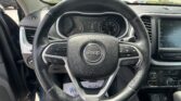 Driver's-seat view of a Jeep steering wheel and dashboard, with a dealership lot and parked cars visible through the windshield àcross the top of the image