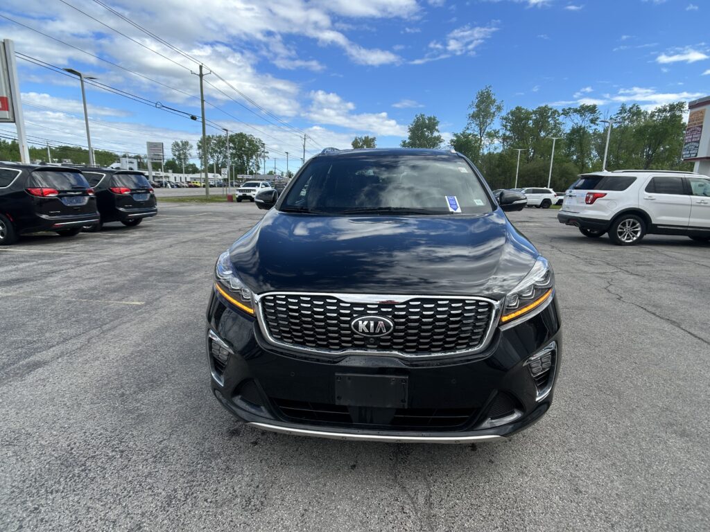 Front view of a dark blue Kia SUV in a busy parking lot on a sunny day, with other cars nearby and power lines above.