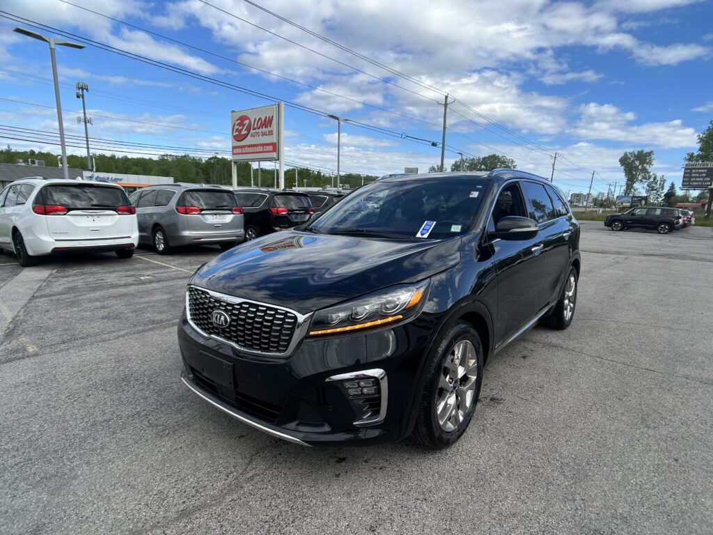 Black Kia SUV in the foreground at a car dealership lot, with white and silver minivans parked behind and a red EZ LOAN sign in the background under a blue sky.