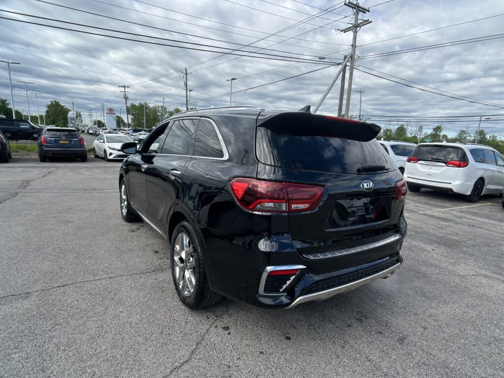 Rear three-quarter view of a black Kia SUV parked in a busy car lot with other vehicles nearby and cloudy skies above.