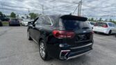 Rear three-quarter view of a black Kia SUV parked in a busy car lot with other vehicles nearby and cloudy skies above.