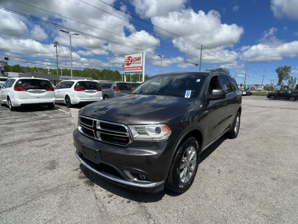 Dark gray Dodge SUV in the foreground at a car dealership lot, with white minivans parked behind and an EZ LOAN Auto Sales sign against a blue sky with clouds.
