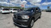 Dark gray Dodge SUV in the foreground at a car dealership lot, with white minivans parked behind and an EZ LOAN Auto Sales sign against a blue sky with clouds.