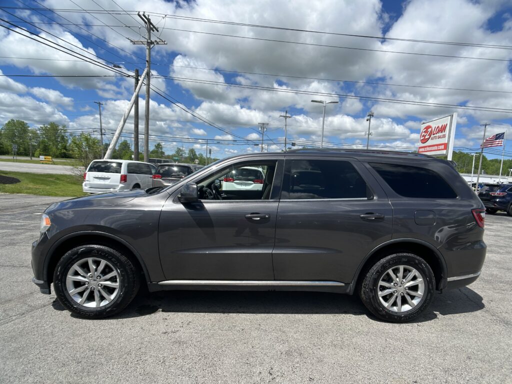 Gray SUV in a car dealership lot with a large E-Z Loan Auto Sales sign and an American flag nearby under a cloudy blue sky and power lines overhead.