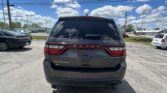 Rear view of a dark gray Dodge Durango SUV in a parking lot under a bright blue sky with scattered clouds and power lines overhead.