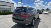 Rear view of a dark gray Dodge Durango SUV in a dealership parking lot with a white building on the left and a blue sky with clouds above