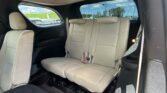 Interior of a vehicle showing beige second-row seats with headrests, black side panels, and a dark floor mat; windows reveal a parking lot outside.