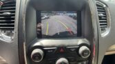 Infotainment screen showing a rearview camera feed with green, yellow, and red guide lines for parking; street with parked cars visible ahead.