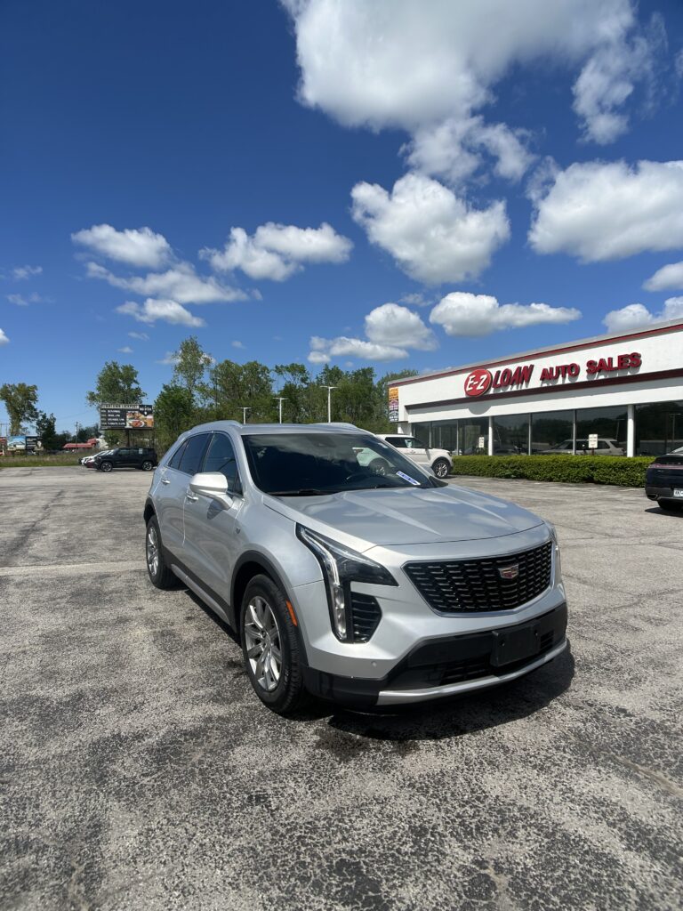 Silver Cadillac SUV parked in a dealership lot in front of EZ Loan Auto Sales, blue sky with fluffy clouds above.