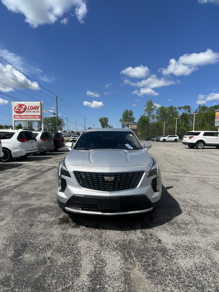 Front view of a silver Cadillac SUV in a crowded car dealership lot with a blue sky above and several white cars lined up nearby, including an E-Z Loan sign in the background.