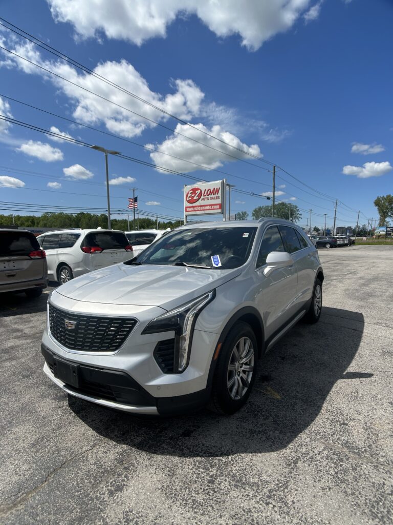 Silver Cadillac SUV parked in a car lot under a bright blue sky with scattered white clouds and an E-Z LOAN Auto Sales sign in the background