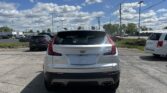 Rear view of a white Cadillac SUV parked in a parking lot, with other cars and a blue sky full of clouds above and power lines crossing the scene.