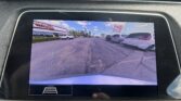 Car infotainment screen displaying rearview camera feed of a parking lot with cars lined up and a dealership sign in the distance