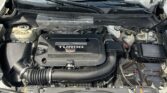 Open car hood revealing the engine bay with a black turbo engine cover labeled TURBO and Cadillac crest; surrounding components include battery on the right, fluid reservoirs, hoses, and wiper area.