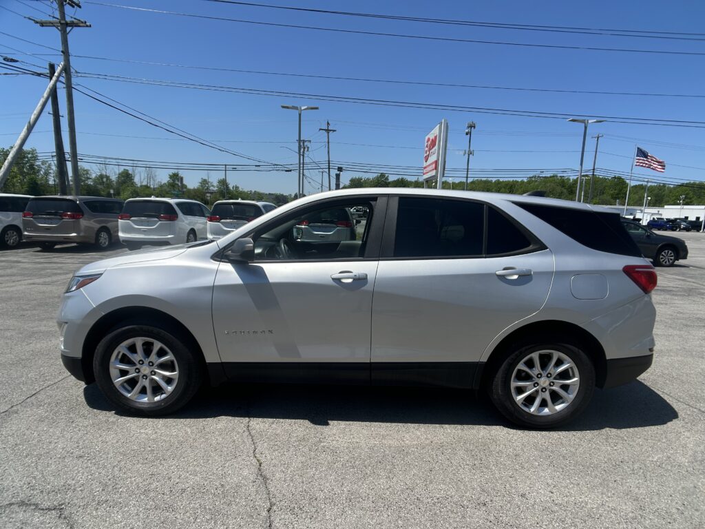 Silver SUV in a car dealership lot with several minivans and an American flag in the background.