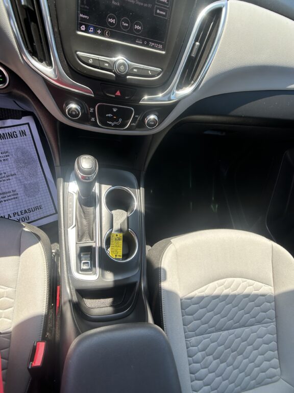 Center console of a car interior featuring a touchscreen infotainment system, climate controls, hazard button, gear shifter, and two cup holders in front of the gear lever.
