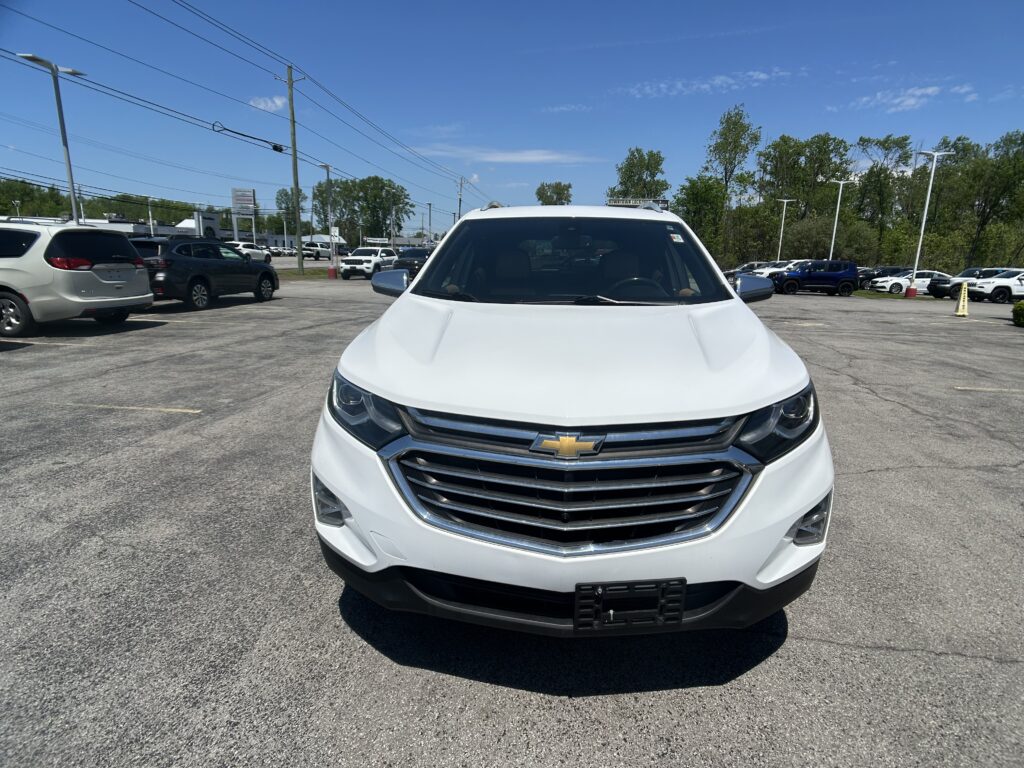 Front view of a white Chevrolet SUV parked in a busy car lot, with other vehicles and power lines in the background under a clear blue sky.