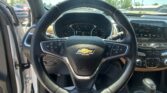 Driver's-eye view of a Chevrolet car interior showing the steering wheel, instrument cluster, and infotainment screen, with a sunny parking lot outside.