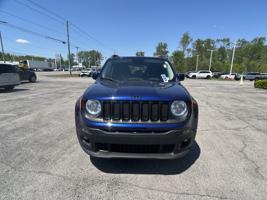 Front view of a blue Jeep Wrangler in a sunny parking lot with other cars and trees in the background.
