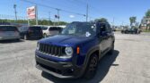 Blue Jeep SUV in a crowded car dealership lot with other cars, signage, and power lines under a clear blue sky.